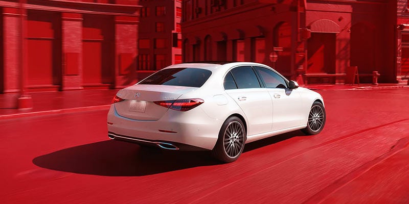 White Mercedes-Benz sedan driving on a city street, viewed from the rear-left angle