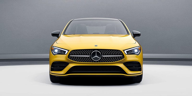 Front view of a shiny yellow Mercedes-Benz with a large, dark grille and bright headlights, centered against a grey background