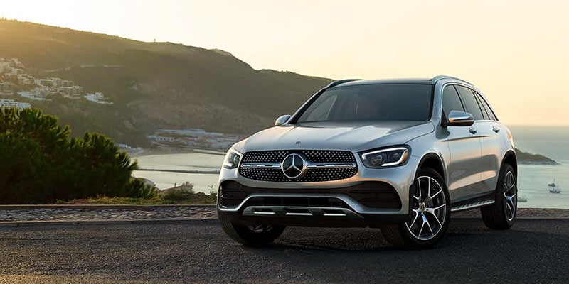 Silver Mercedes-Benz SUV parked on a coastal road with hills and ocean views in the background during sunset