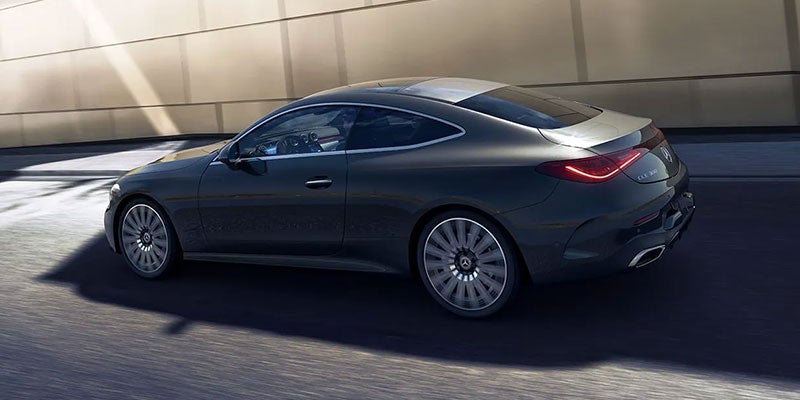 A dark gray Mercedes-Benz luxury coupe driving on a road, seen from the rear three-quarter view