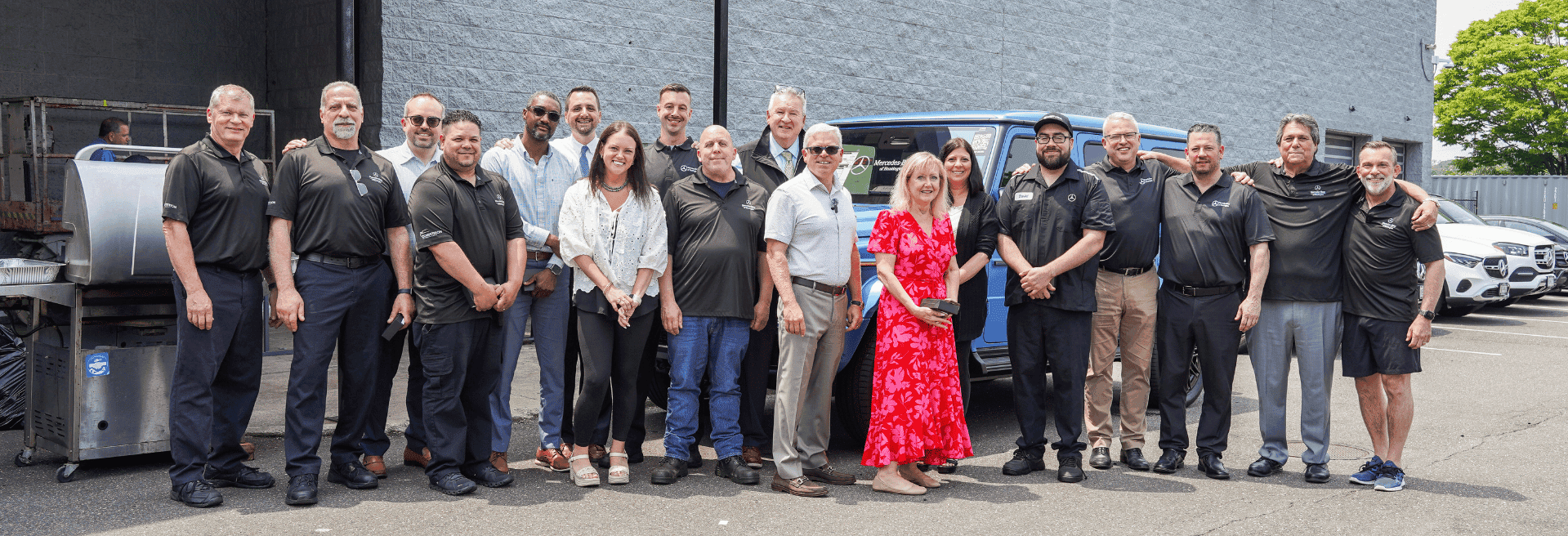 A large group of ~20 people, men and women, pose together for a photo outside a business, with a grill and cars in the background.E