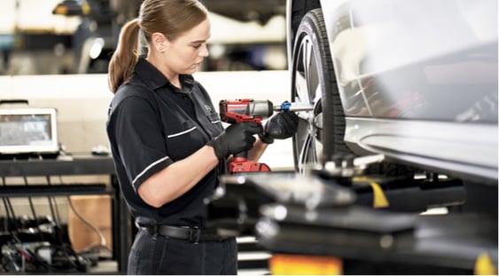 A female mechanic in a black uniform tightens a lug nut on a lifted car's wheel with a power tool.