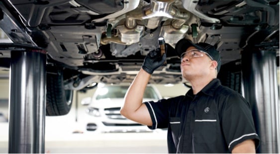 Technician in safety goggles and gloves inspecting the undercarriage of a car on a lift with a flashlight.