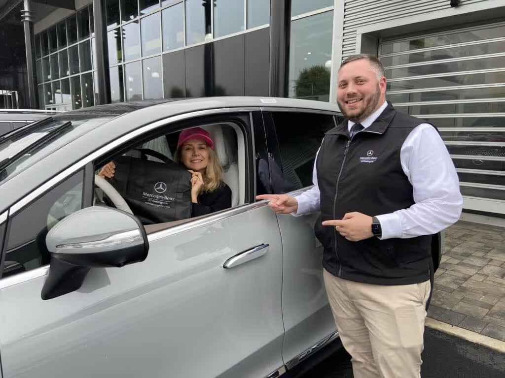 Woman in a Mercedes-Benz holding a Mercedes bag, with a smiling salesman pointing to her.