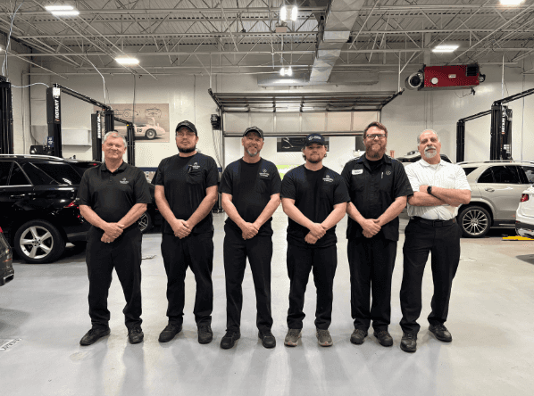 Six men, some wearing black uniform shirts and hats, stand side-by-side in a modern auto garage.