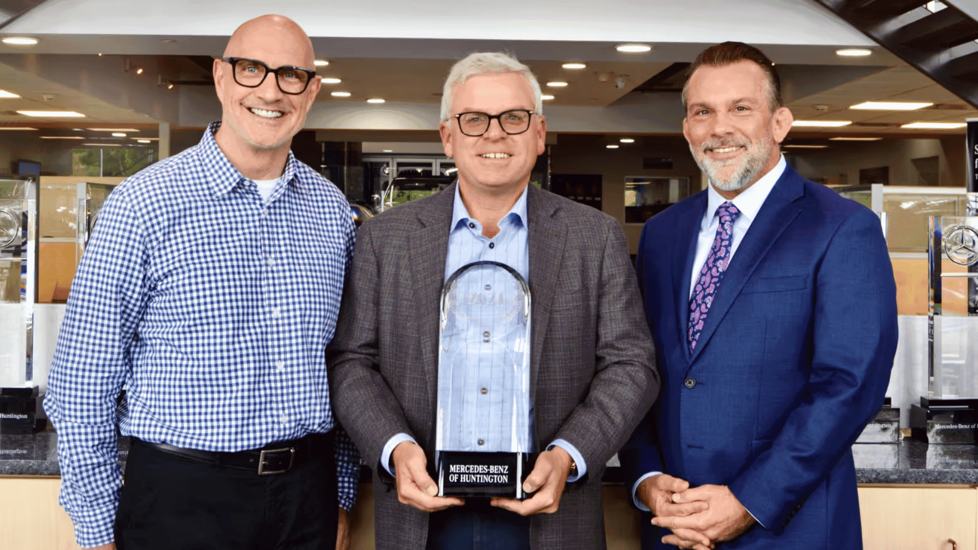 Three men smiling; the middle man holds a clear Mercedes-Benz of Huntington award with a black base.