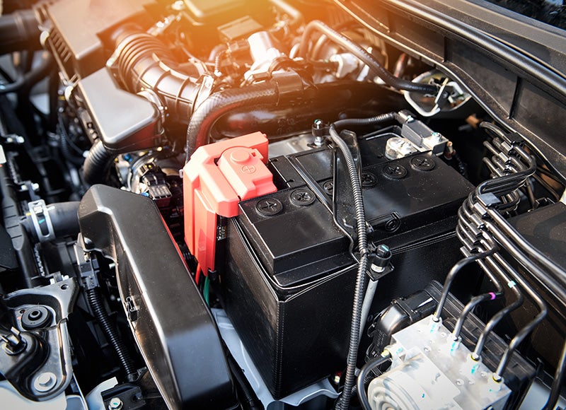 Close-up of a car engine compartment showing a black automotive battery with a red positive terminal cover and surrounding engine components